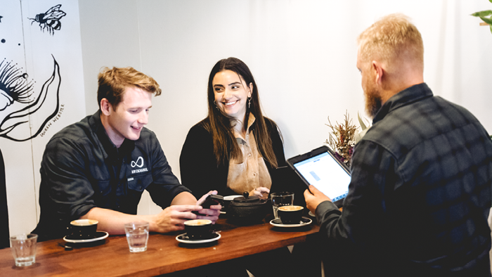 three people sitting in a cafe smiling and looking at devices