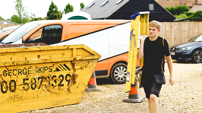 young man carrying step ladder on shoulders walking past a large skip bin