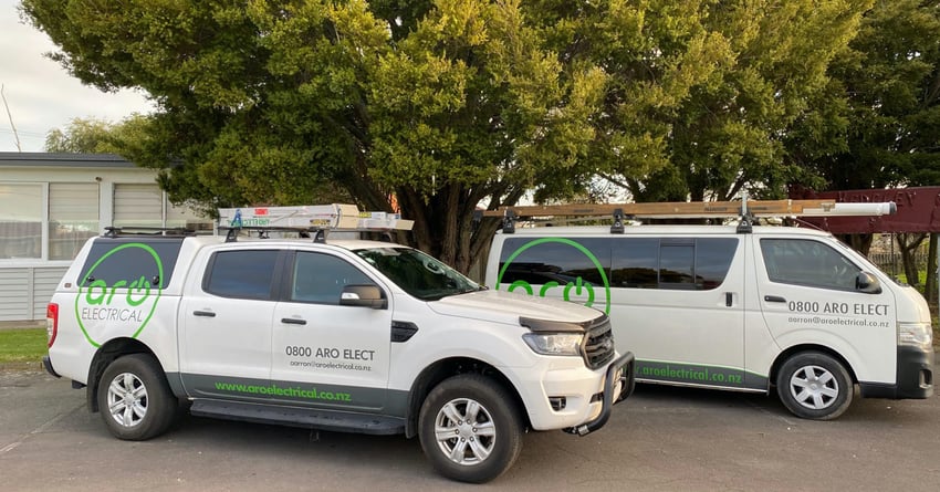 The Aro Electrical fleet, a white ute and a white van, parked together in front of a school building