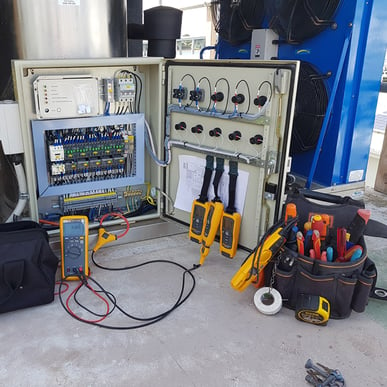 A photo of the inside of an electrical cabinet hooked up to testing equipment. Next to it sits a very full tool bag.
