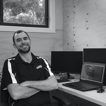 A black and white photo of electrician, Kiah, smiling with arms crossed and sitting in his office in front of a laptop.