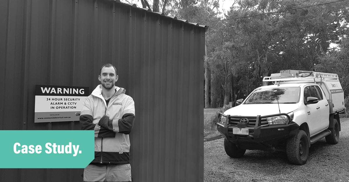A black and white photo of electrician and tradify customer Kiah smiling and standing with arms crossed in front of a dark coloured shed