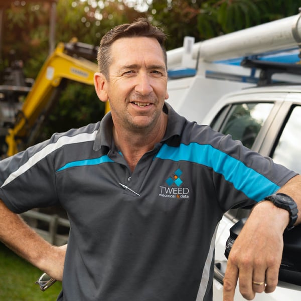 Michael from Tweed Electrical leans against a UTE, one hand, holding glasses, resting on his hip. There is a cool yellow digger in the background.