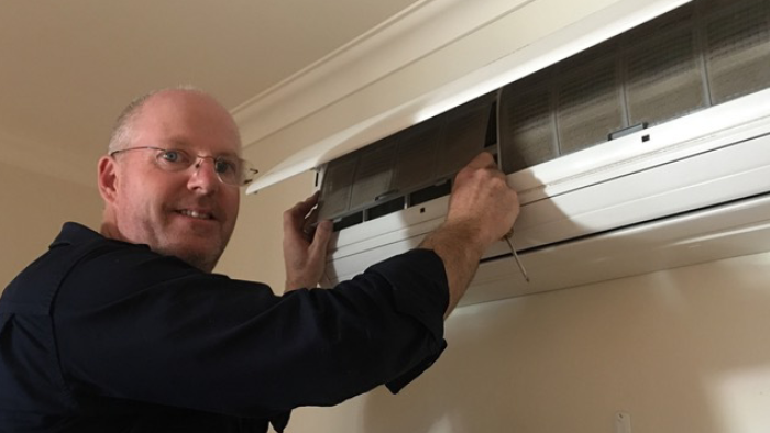 greg begelhole looking at camera while standing in-front of a wall mounted heat pump 