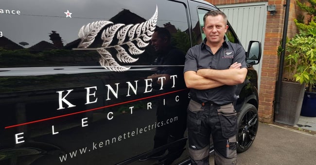 An electrician standing in front of his black with a sign for Kennett Electrics and a silver fern emblem