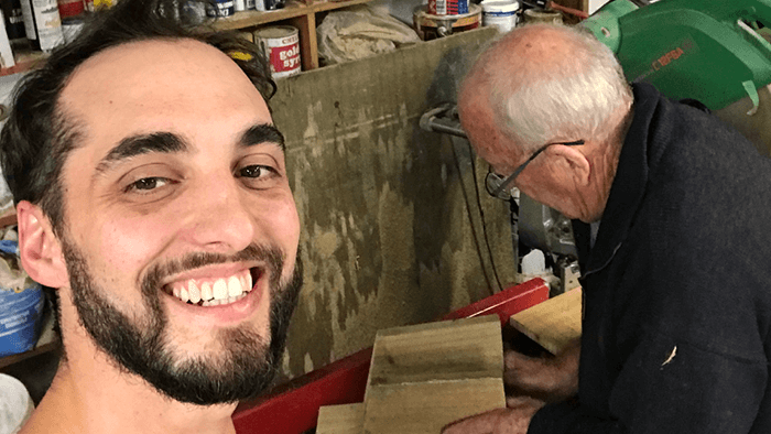 Mat_Leandro_selfie of a young man looking at the camera smiling while older man is working on wooden frames 