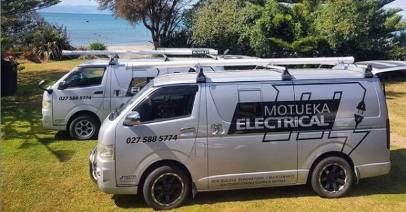 Two silver Motueka Electrical vans parked up on the grass next to a sandy beach