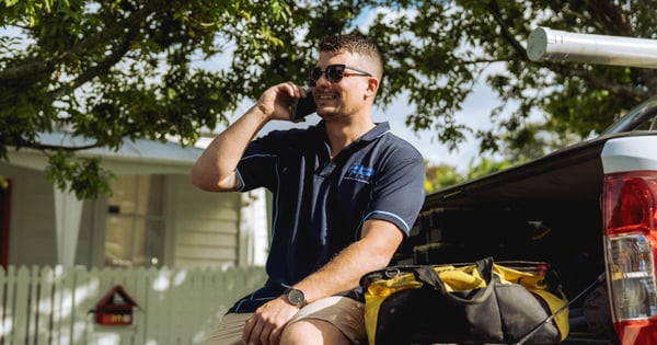 Plumber sitting on edge of car speaking on phone-min